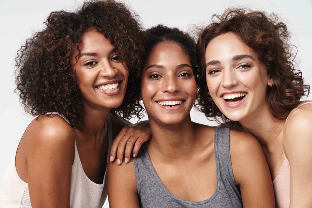 Three smiling women posing together, showcasing healthy, glowing skin and diverse complexions.